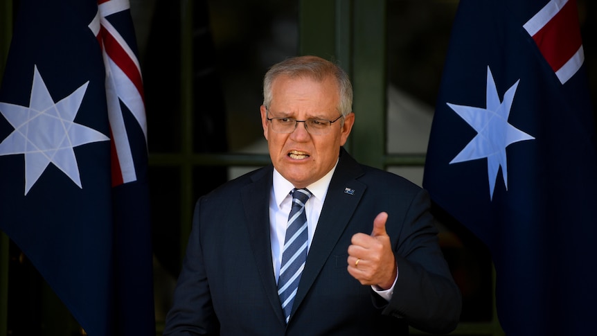 Grey-haired man in glass and blue striped tie, blue jacket gestures with thumb