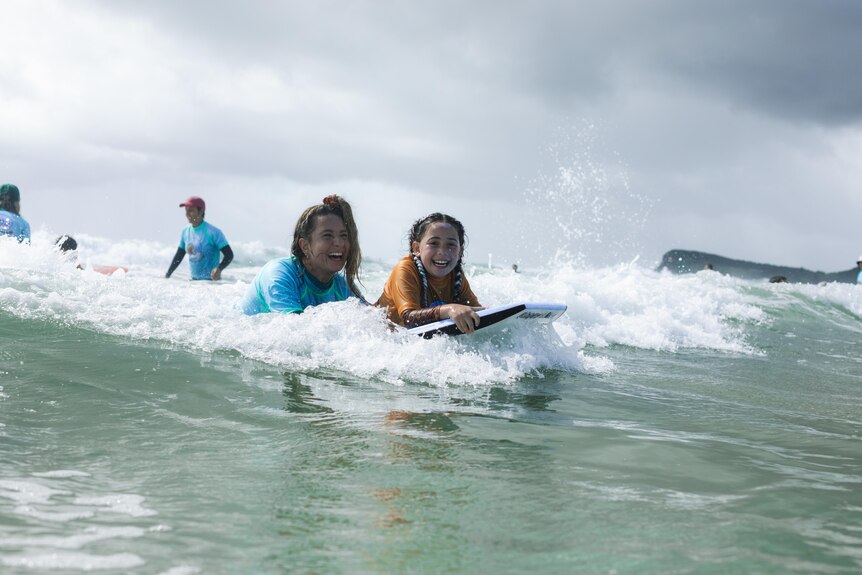 A young girl with an orange rashie, boogie boarding with a volunteer wearing blue. Both smiling and laughing