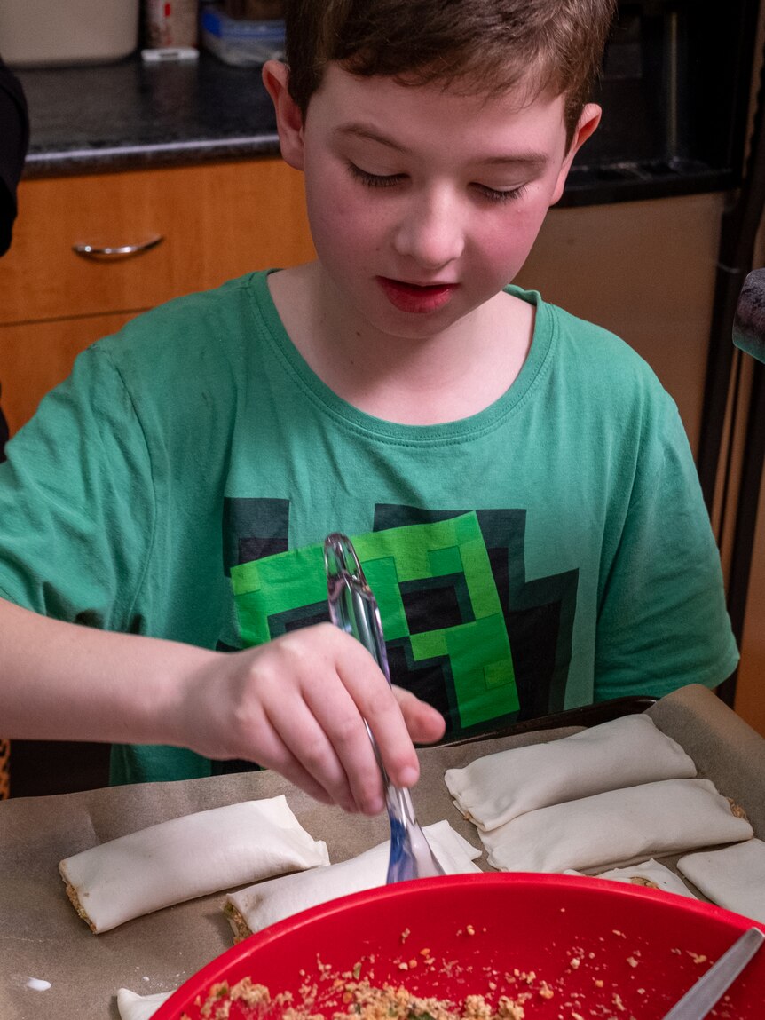 A little boy makes sausage rolls in his kitchen at home.