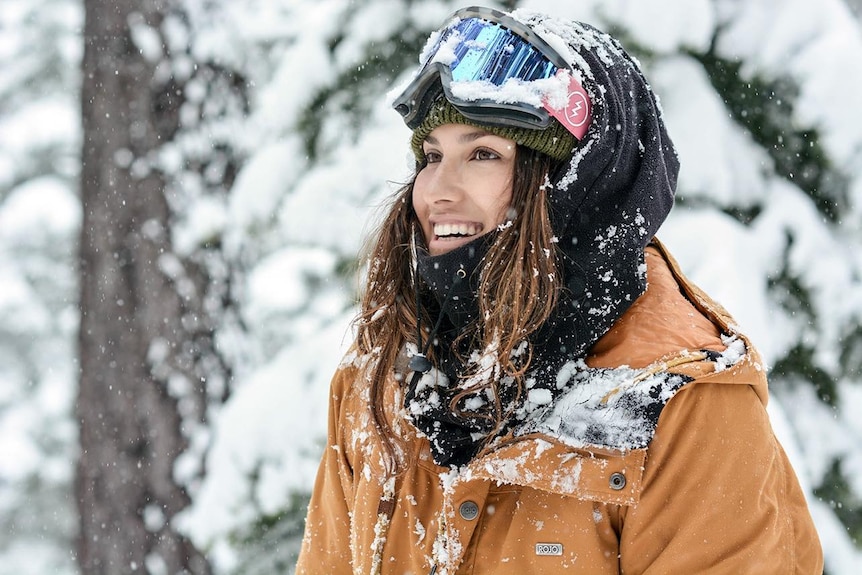 Rani smiles and looks away from the camera to the left while wearing snow gear and standing amongst trees and snow falls.