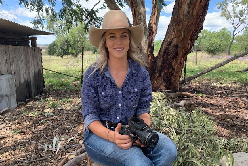 A young woman in sits in a backyard holding a camera.