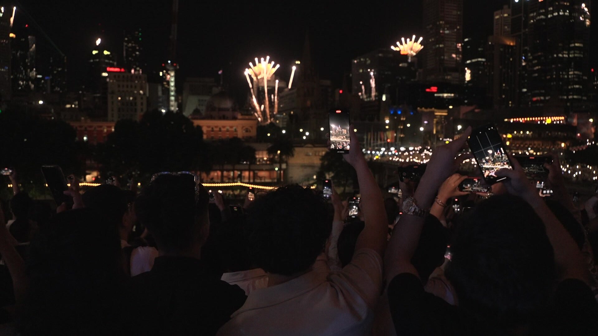 Uma foto tirada atrás de uma multidão, muitos dos quais seguram seus telefones filmando fogos de artifício vindos de prédios próximos da cidade à noite.