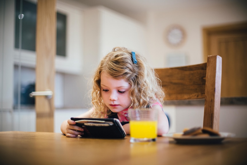 Little girl with blonde hair sits at table looking at phone