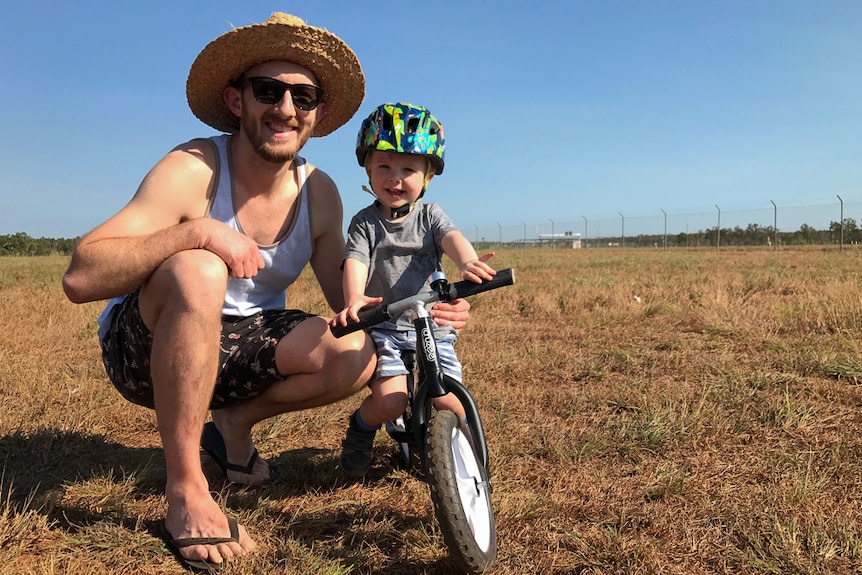 A man in a straw hat and sunglasses crouches next to his son who is straddling a balance-bike on a field of dry grass.