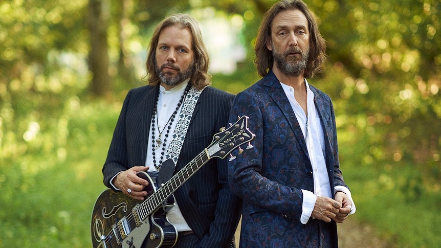 Chris and Rich Robinson of The Black Crowes stand outside wearing suits. Rich holds a guitar and wears beads. Both have beards.