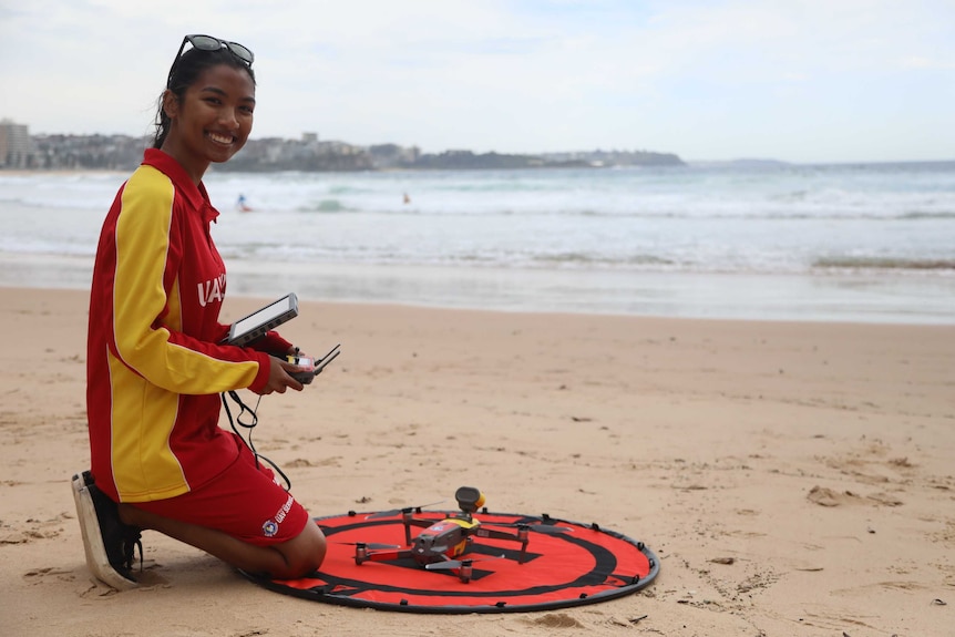 A girl in a surf life saving uniform kneels in front of a drone on the sand at a beach