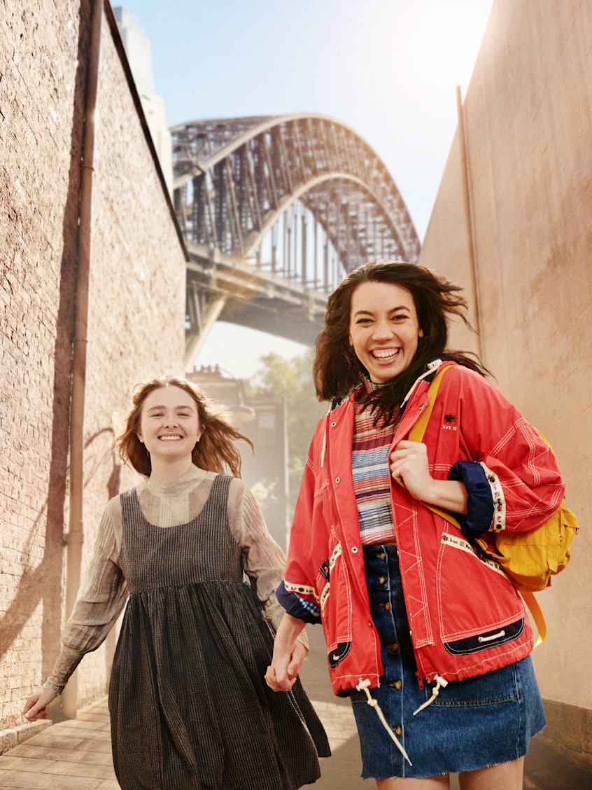 Two young women holding hands and smiling run down a brick-lined laneway, with Sydney Harbour Bridge seen in background.