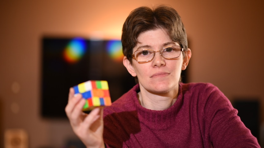 A young white woman with short brown hair and glasses. She's wearing a maroon jumper and holding a rubix cube