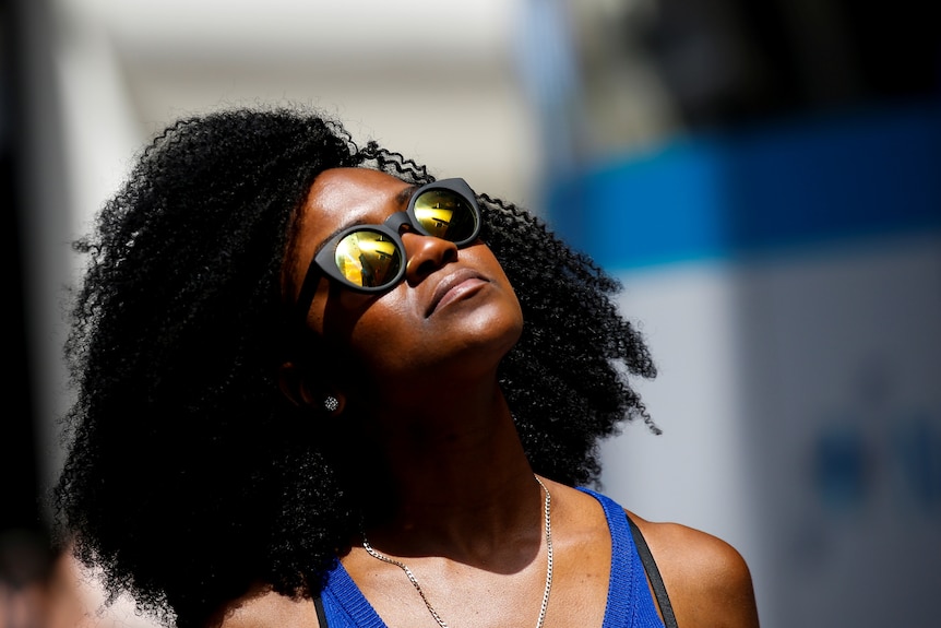 A woman in reflective sunglasses with dark curly hair looks up at the sun 