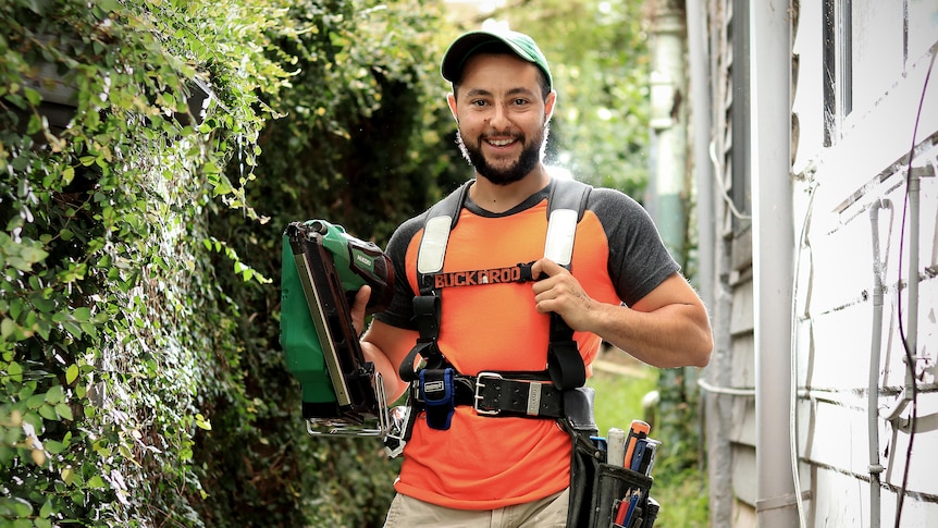 A bearded man stands outside a house with his tool belt and wearing a green hat with orange and grey shirt.