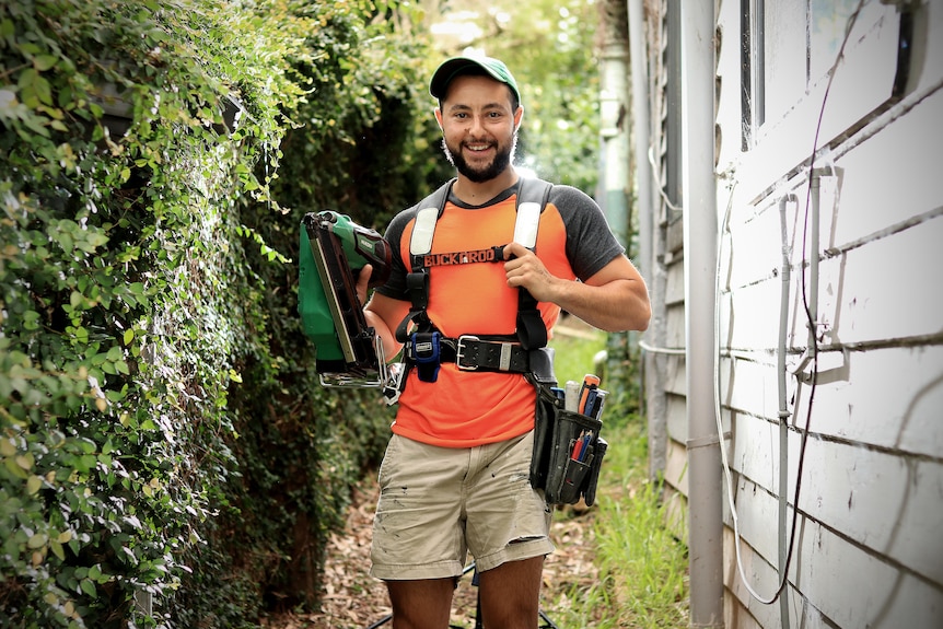 A bearded man stands outside a house with his tool belt and wearing a green hat with orange and grey shirt.
