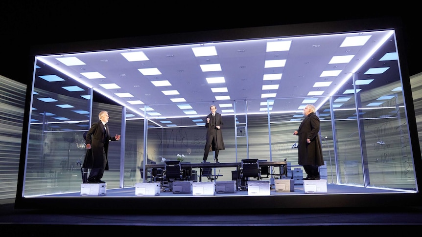 In a glass box on stage, two men stand on document boxes and one stands on a boardroom desk.