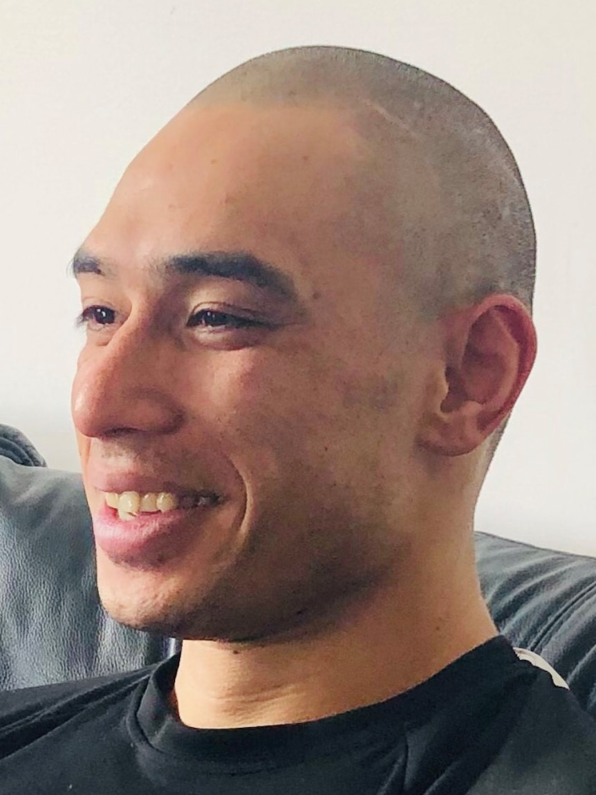A profile shot of a young man in a black t-shirt with a bald head, smiling.