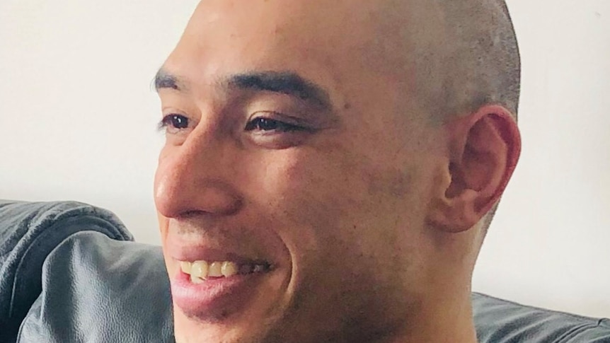 A profile shot of a young man in a black t-shirt with a bald head, smiling.