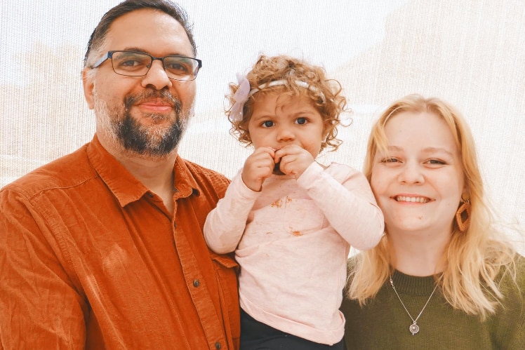 A young family. The man man is wearing glasses, the lady has blonde hair, and they're holding a young child