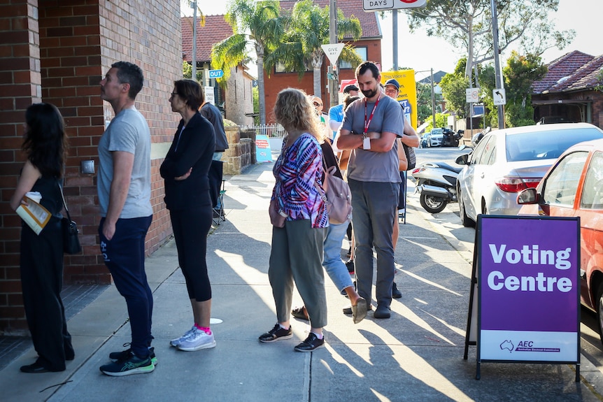 People lining up on a footpath to enter a voting centre. Cars parked nearby, and a Voting Centre sign on the footpath