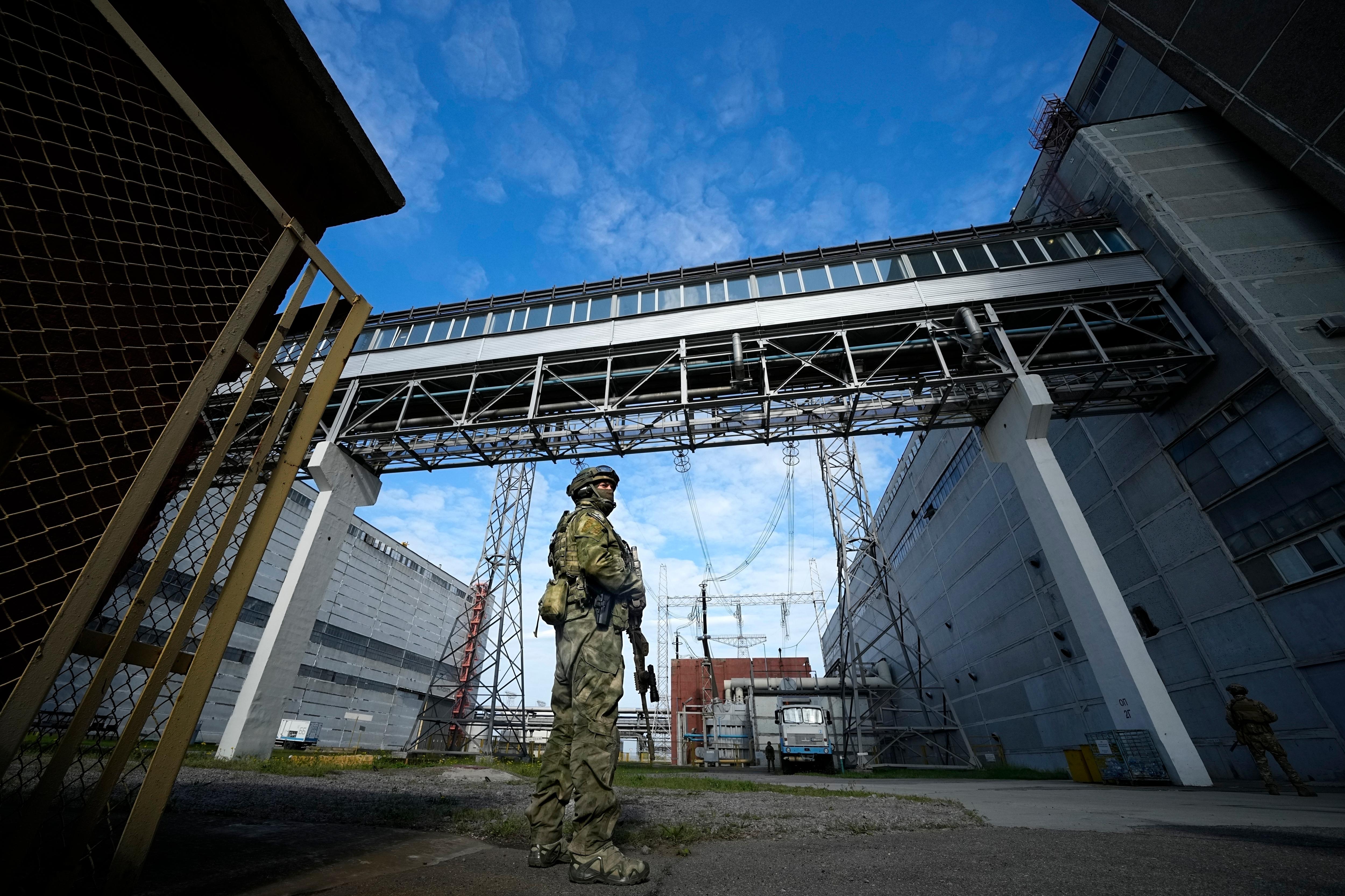 A Russian soldier in full combat gear holds a weapon as he stands between two industrial buildings near power lines.