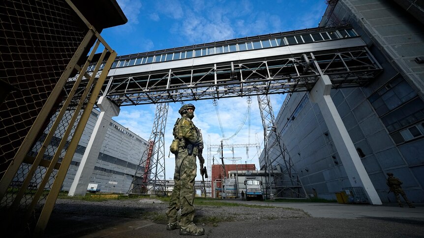 A Russian soldier in full combat gear holds a weapon as he stands between two industrial buildings near power lines.