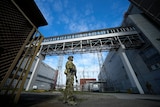 A Russian soldier in full combat gear holds a weapon as he stands between two industrial buildings near power lines.