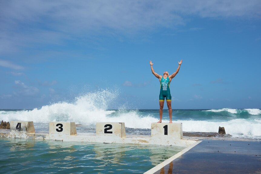 Katie Kelly lifts her arms to the sky while standing on the edge of an ocean pool.