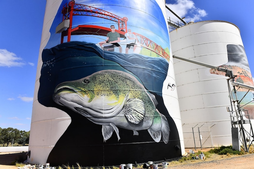A large silo with a large face framing blue water with a giant Murray cod, two men fishing and a big red bridge.