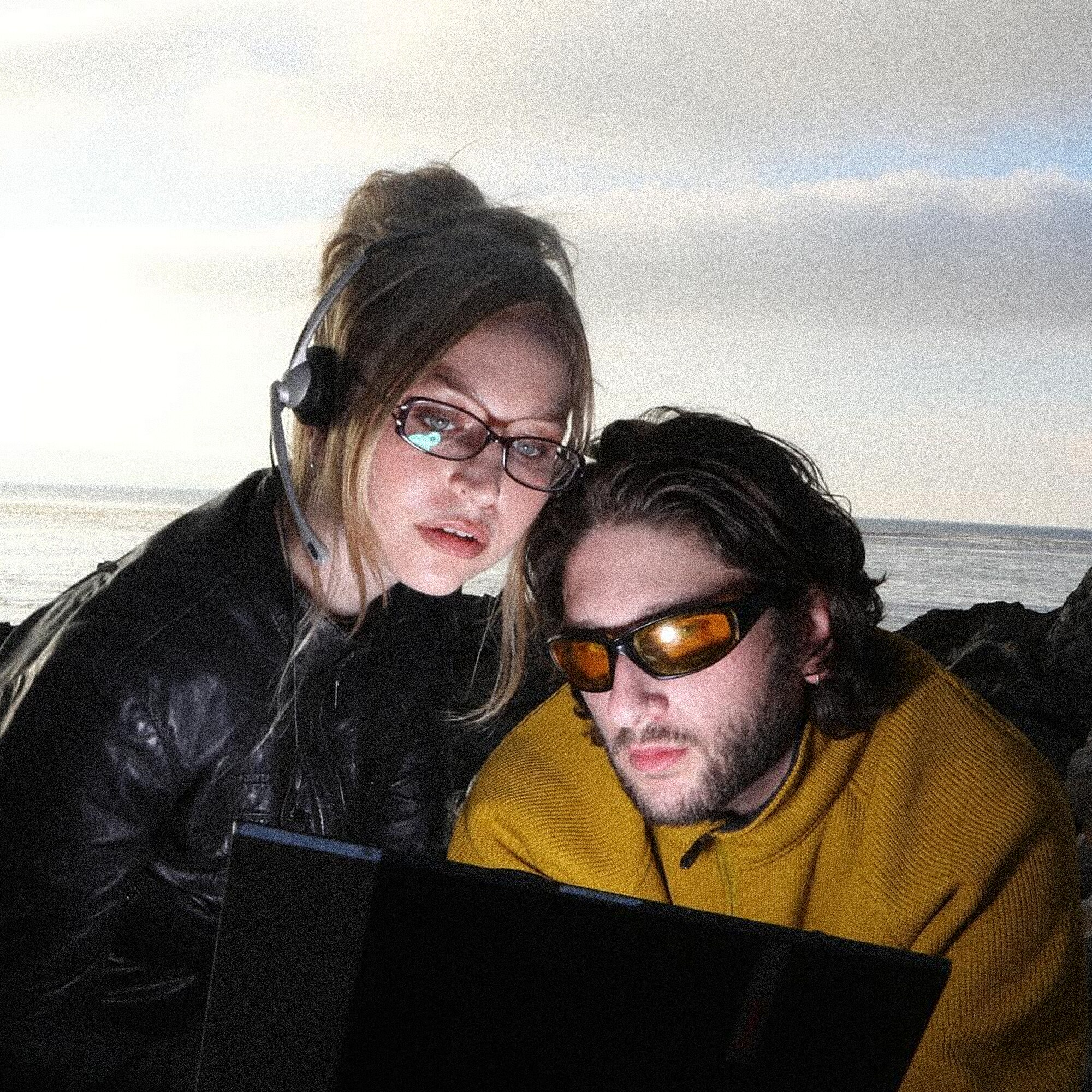 A man and woman with sunglasses on looking at a laptop screen. The background behind them is the ocean and sky. 