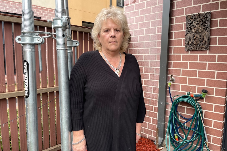 A woman with a blank face stands outside an apartment.