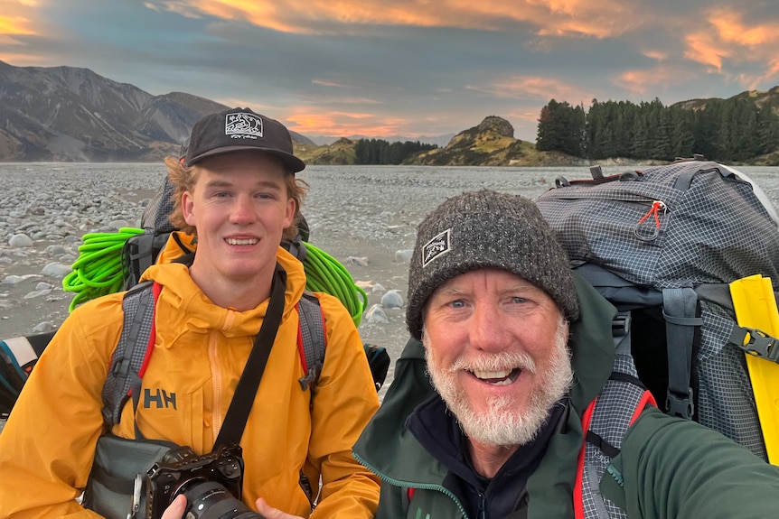 A young man and his middle-aged father pose for a selfie, wearing outdoorsy camping gear carrying backpacks.
