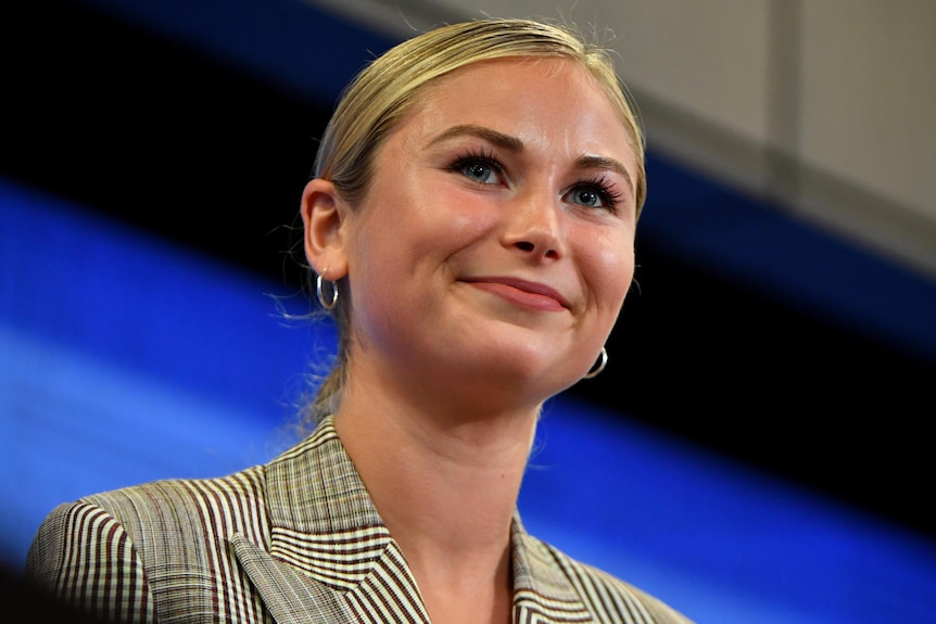 A close up of a woman smiling towards the audience. Her blonde hair is pulled back and she's wearing a plaid jacket.