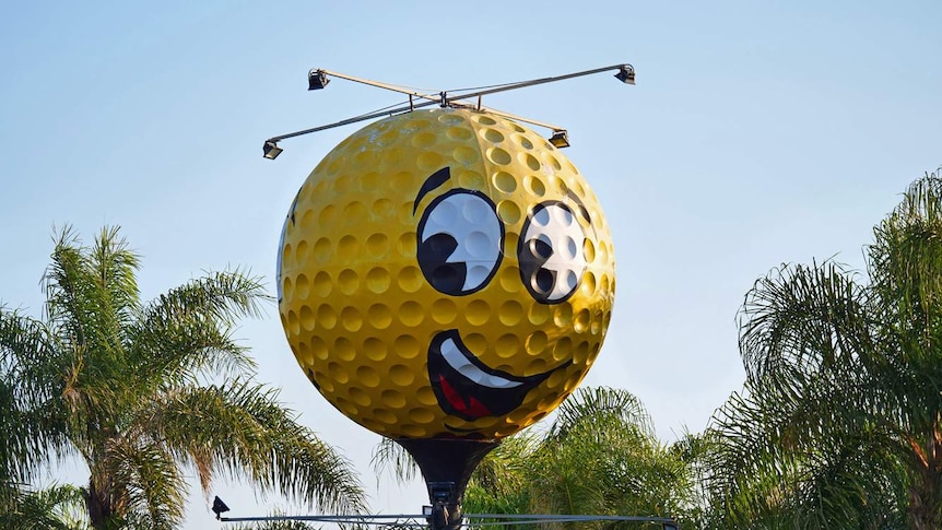 An oversized, yellow golf ball with a smiling face painted on it towers over palm trees.