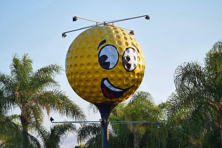 An oversized, yellow golf ball with a smiling face painted on it towers over palm trees.