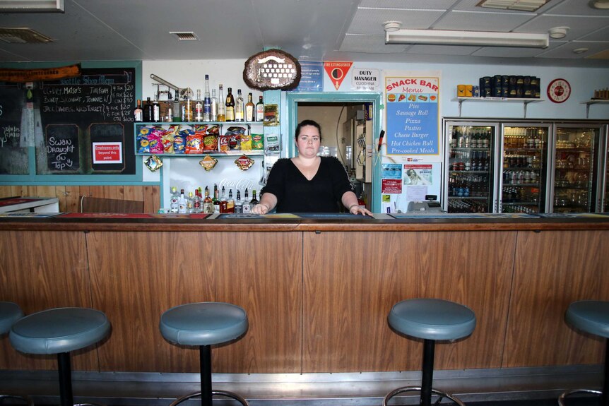 Cliodhna stands behind the bar, with liquor and a beer fridge in the background, trophies on the wall, and a specials board.
