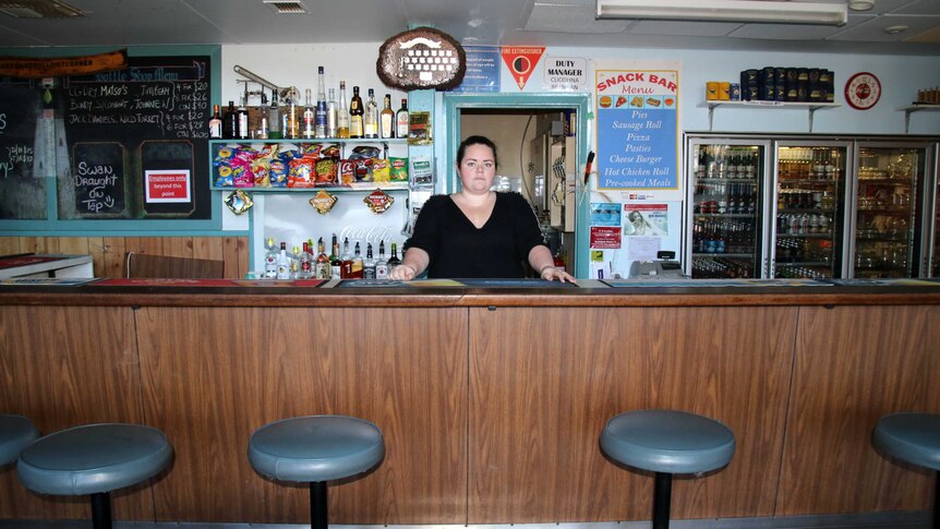 Cliodhna stands behind the bar, with liquor and a beer fridge in the background, trophies on the wall, and a specials board.