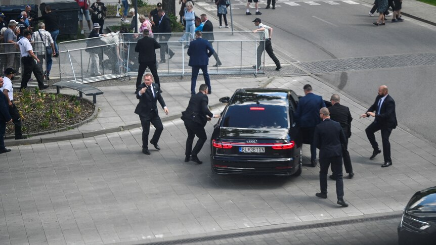 Security guards in black suits rush around a black parked car as members of the public look on