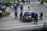 Security guards in black suits rush around a black parked car as members of the public look on