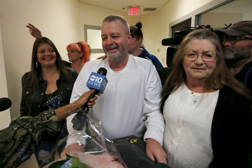 a man with a beard and a white shirt has a microphone infront of him and a news camera beside him along with a group of women