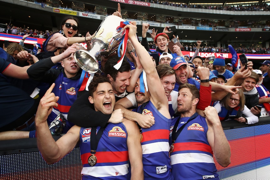 A group of excited AFL players pose with and point to the premiership cup, as delighted supporters hug them from behind.