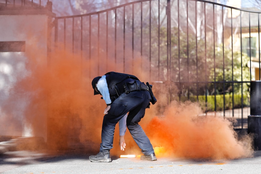 A police officer bends down to pick up a flare releasing orange smoke outside The Lodge