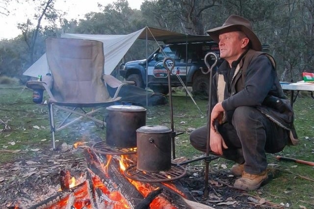 Tim Bates squats in front of a campfire with his tent and 4WD in the background
