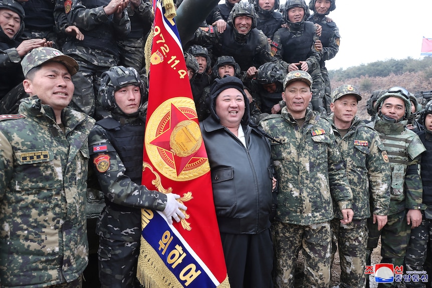 Kim Jong Un smiles as he stands with soldiers in uniform next to a flag.