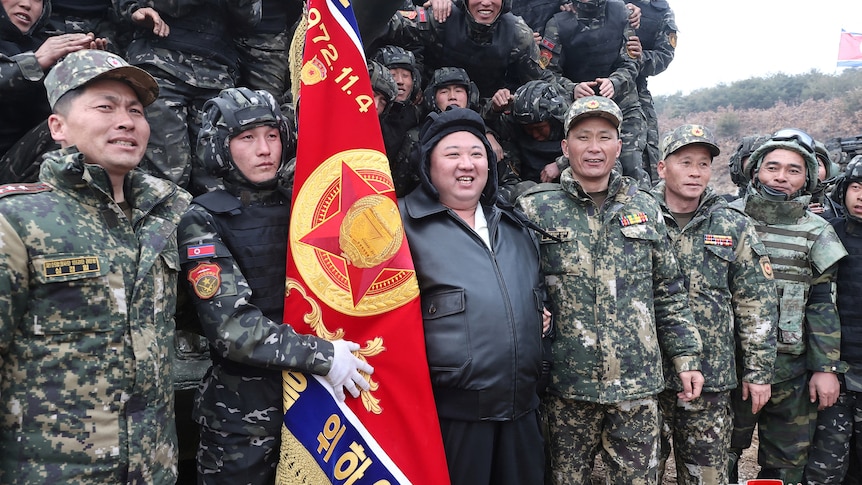 Kim Jong Un smiles as he stands with soldiers in uniform next to a flag.