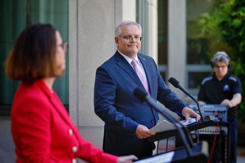 Scott Morrison looks at Anne Ruston as she addresses the media at a lectern in a courtyard