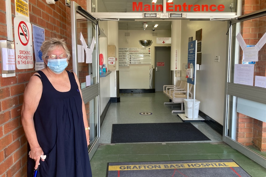 woman holding walking stick standing outside hospital door with door mat reading Grafton Base Hospital