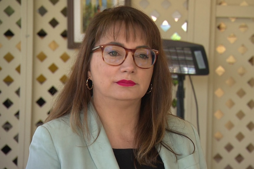 A woman with long brown hair and red glasses looks slightly off camera with a serious expression