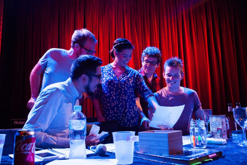 A group of comedians recording the Dragon Friends podcast on a stage with red curtains in the background