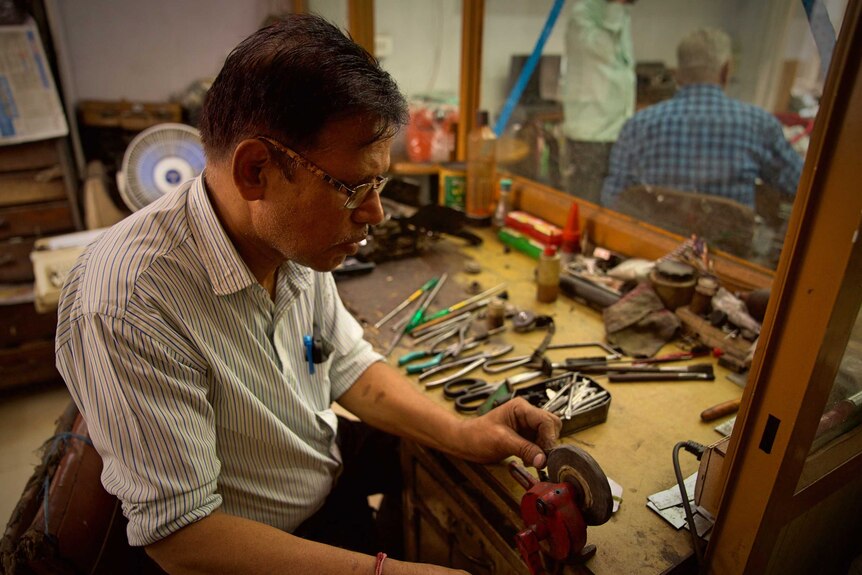 A man works at a desk covered in tools, next to a small fan.