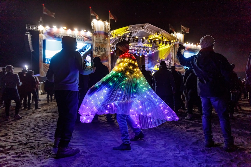 A person in a light-up cape dances at night in front of a stage among a crowd of people