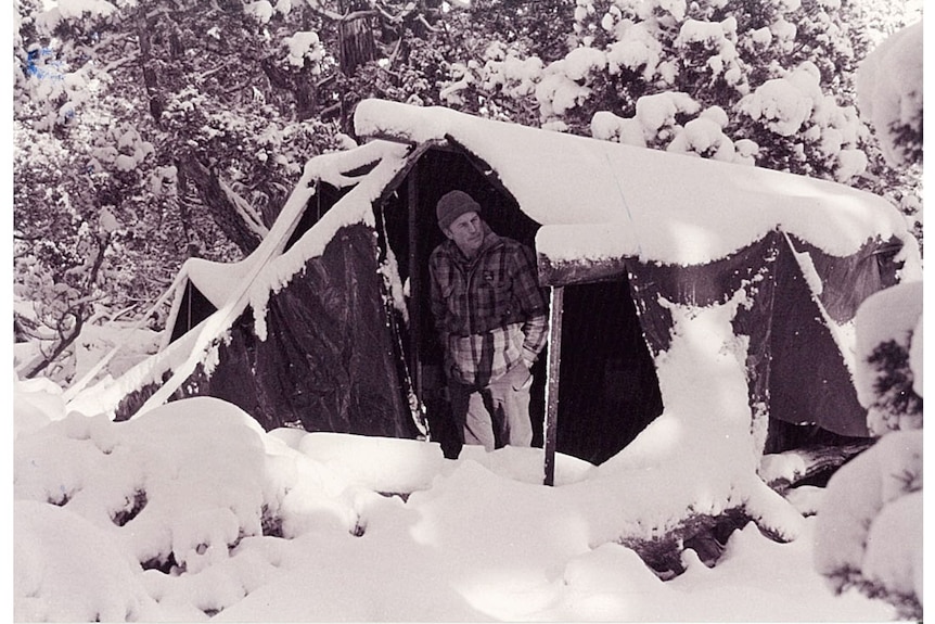 A man pokes his head out of a tent that has been heavily covered in snow at a snowy wilderness campsite.