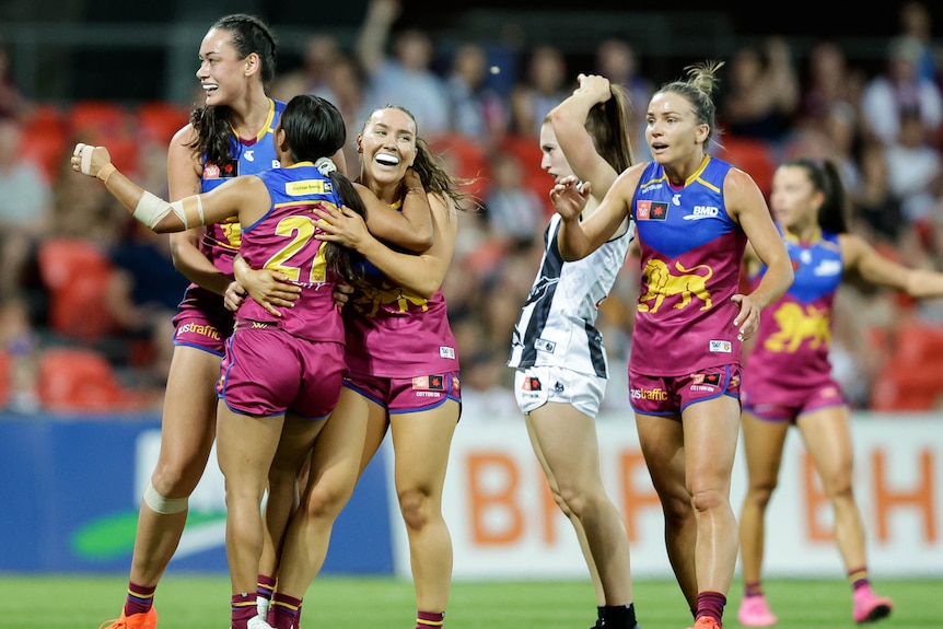 Un grupo de jugadores de los Lions celebra un gol mientras un jugador de Collingwood parece decepcionado detrás de ellos.