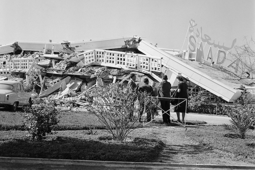 A black and white image shows a crumpled building and people walking in the foreground. 
